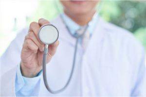 em primeiro plano é possível observar a mão de um médico com um estetoscópio e ao fundo seu corpo usando avental branco por cima de gravata e camisa azul claro