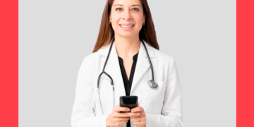 mulher de jaleco branco e aparelho de ouvir batimentos cardíacos segurando um celular nas mãos e sorri para a câmera