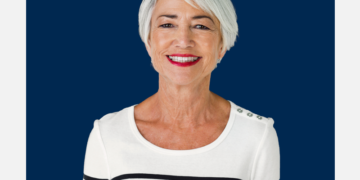 Mulher de cabelo grisalho e curto sorri para a câmera sob um fundo azul marinho e branco.