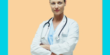 Médica de jaleco e braços cruzados olha pra câmera sobre um fundo bege e azul