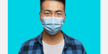 Homem oriental de máscara cirúrgica olha para a câmera sob um fundo branco e azul