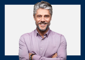 Homem de meia idade sorri para câmera com braços cruzados sob um fundo cinza e azul marinho.