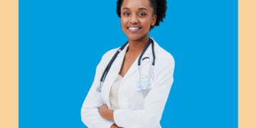 Médica olha para a câmera de frente de braços cruzados sobre fundo azul e bege.