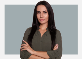 Mulher branca com cabelos lisos e pretos olha para a câmera com oa braços cruzados sob um fundo cinza e branco