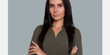 Mulher branca com cabelos lisos e pretos olha para a câmera com oa braços cruzados sob um fundo cinza e branco