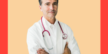 Médico branco olha para a câmera de braços cruzados sob um fundo bege e coral.