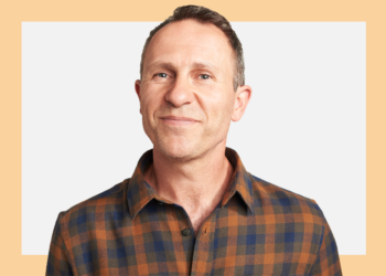 Homem de camisa xadrez sorri para a câmera sob um fundo bege e cinza.