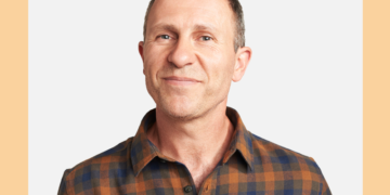 Homem de camisa xadrez sorri para a câmera sob um fundo bege e cinza.