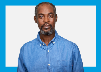 Homem negro de camisa azul jeans de frente e semblante sério sob um fundo azul e branco.