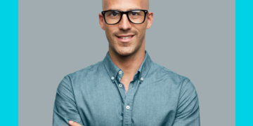 Homem olha e sorri para a câmera de braços cruzados sobre fundo cinza e azul