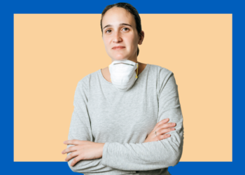 Mulher com máscara pendurada no pescoço sorri levemente com os braços cruzados olhando para a câmera.