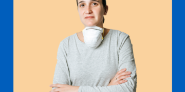 Mulher com máscara pendurada no pescoço sorri levemente com os braços cruzados olhando para a câmera.