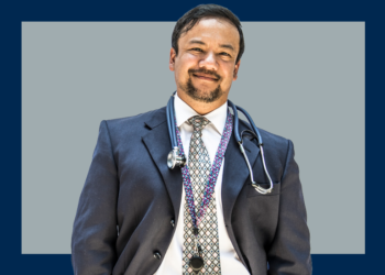 Médico de terno e estetoscópio olha de frente para a câmera sobre um fundo cinza e marinho