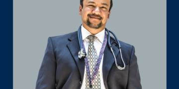 Médico de terno e estetoscópio olha de frente para a câmera sobre um fundo cinza e marinho