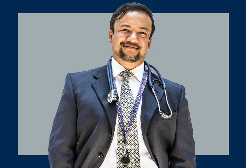 Médico de terno e estetoscópio olha de frente para a câmera sobre um fundo cinza e marinho