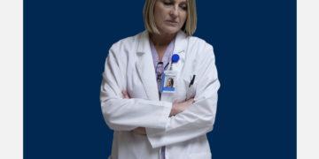 Médica olha pra baixo, cabisbaixa, sobre um fundo branco e azul marinho