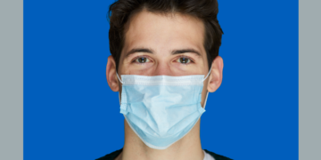 Homem de máscara cirúrgica olha pra câmera sobre fundo cinza e azul