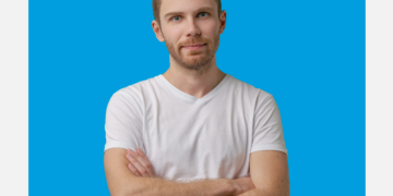 Homem de frente, braços cruzados e camiseta branca olha para a câmera com semblante sério.