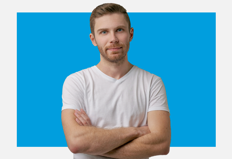 Homem de frente, braços cruzados e camiseta branca olha para a câmera com semblante sério.