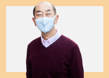 Homem de máscara cirúrgica e blusa vinho olha para a câmera sobre um fundo branco e bege