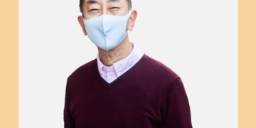 Homem de máscara cirúrgica e blusa vinho olha para a câmera sobre um fundo branco e bege