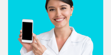 Mulher com cabelos presos e jaleco segura celular sorrindo sobre um fundo azul e branco.