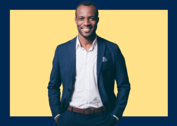 Homem de camisa branca e paletó azul marinho sorri, com as mãos nos bolsos, sobre um fundo amarelo e azul marinho.