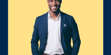 Homem de camisa branca e paletó azul marinho sorri, com as mãos nos bolsos, sobre um fundo amarelo e azul marinho.