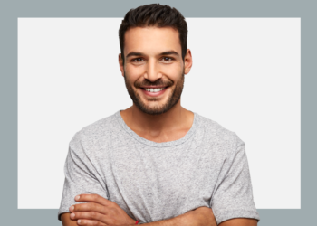 Homem de camisa cinza e braços cruzados olha para a câmera sorrindo sobre um fundo branco e cinza.