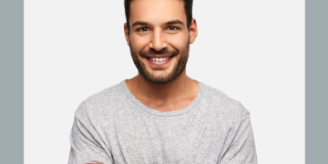 Homem de camisa cinza e braços cruzados olha para a câmera sorrindo sobre um fundo branco e cinza.