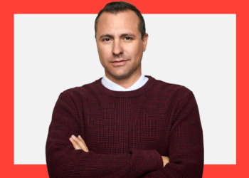 Homem de blusa vinho e braços cruzados olha com semblante sério para a câmera sobre um fundo branco e coral.