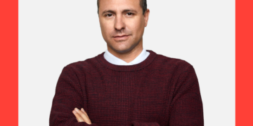 Homem de blusa vinho e braços cruzados olha com semblante sério para a câmera sobre um fundo branco e coral.