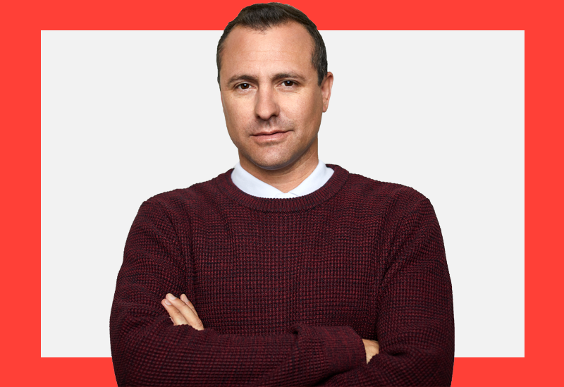 Homem de blusa vinho e braços cruzados olha com semblante sério para a câmera sobre um fundo branco e coral.