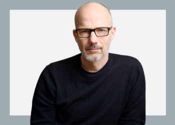 Homem de óculos e blusa preta olha com semblante sério para a câmera sobre fundo branco e cinza