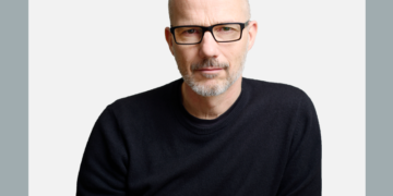 Homem de óculos e blusa preta olha com semblante sério para a câmera sobre fundo branco e cinza