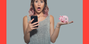 Mulher de cabelos rosas e blusa cinza segura o celular numa mão e um cofrinho em formato de porquinho na outra, fazendo cara de espanto. O fundo é cinza e coral.