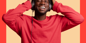 Homem de blusa vermelha e de fones de ouvidos sorri de olhos fechados sobre um fundo bege e coral.