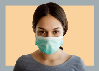 Mulher de máscara cirúrgica olha para a câmera sobre um fundo cinza e bege.