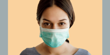 Mulher de máscara cirúrgica olha para a câmera sobre um fundo cinza e bege.