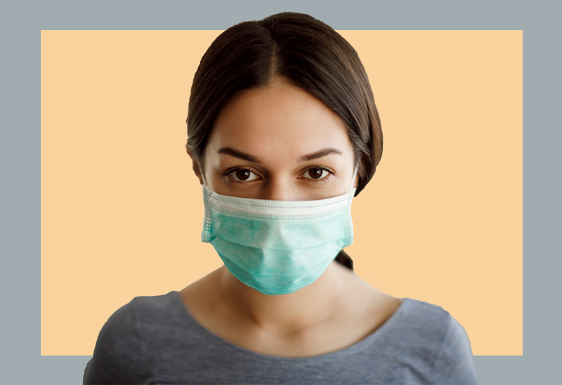 Mulher de máscara cirúrgica olha para a câmera sobre um fundo cinza e bege.