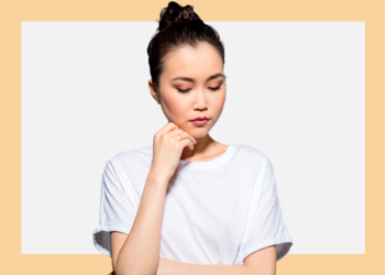 Mulher de cabelos presos e camiseta branca olha para baixo com a mão no queixo sobre um fundo branco e bege.