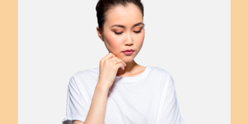 Mulher de cabelos presos e camiseta branca olha para baixo com a mão no queixo sobre um fundo branco e bege.